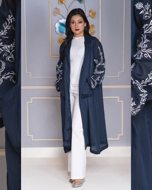 Woman wearing a navy blue embroidered coat over a white outfit against a decorative wall.