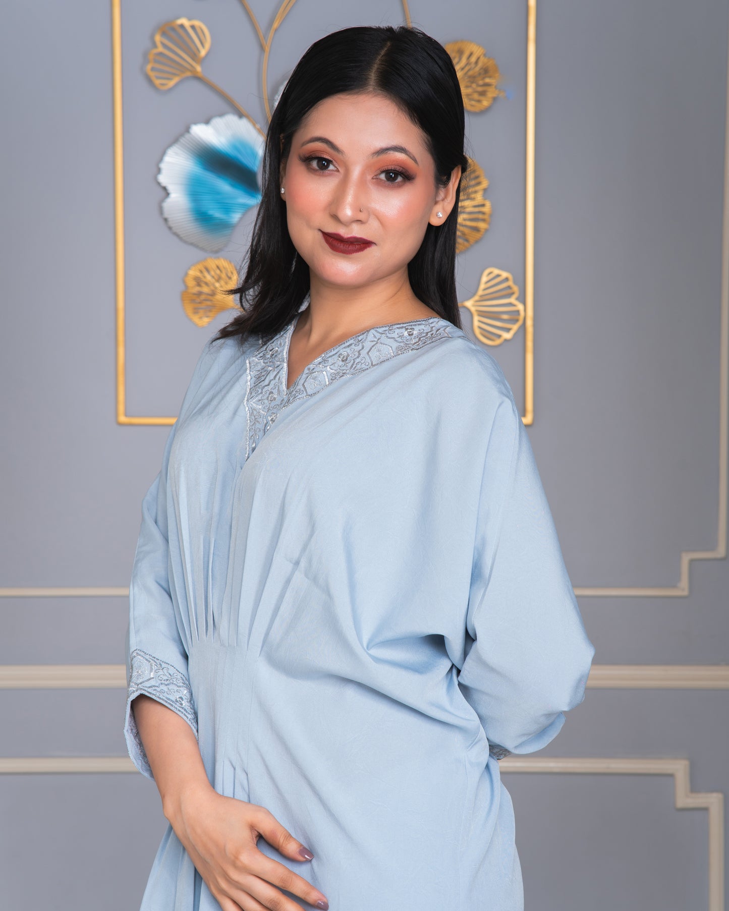 Woman wearing a light blue dress with decorative trim against a gray wall with gold accents.