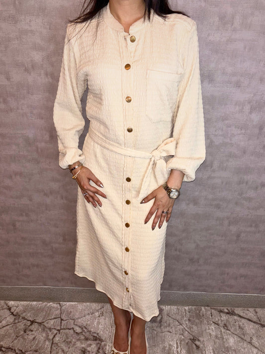 Experience versatile elegance with the Gilded Ivory Dress, a highlight of Taraashi's Platinum Collection. Crafted from uniquely textured fabric in ivory, this versatile piece blends everyday comfort with understated polish. Styled like a classic shirt dress with a defining tie belt, it's distinguished by a row of luminous gold buttons lending an elevated touch.