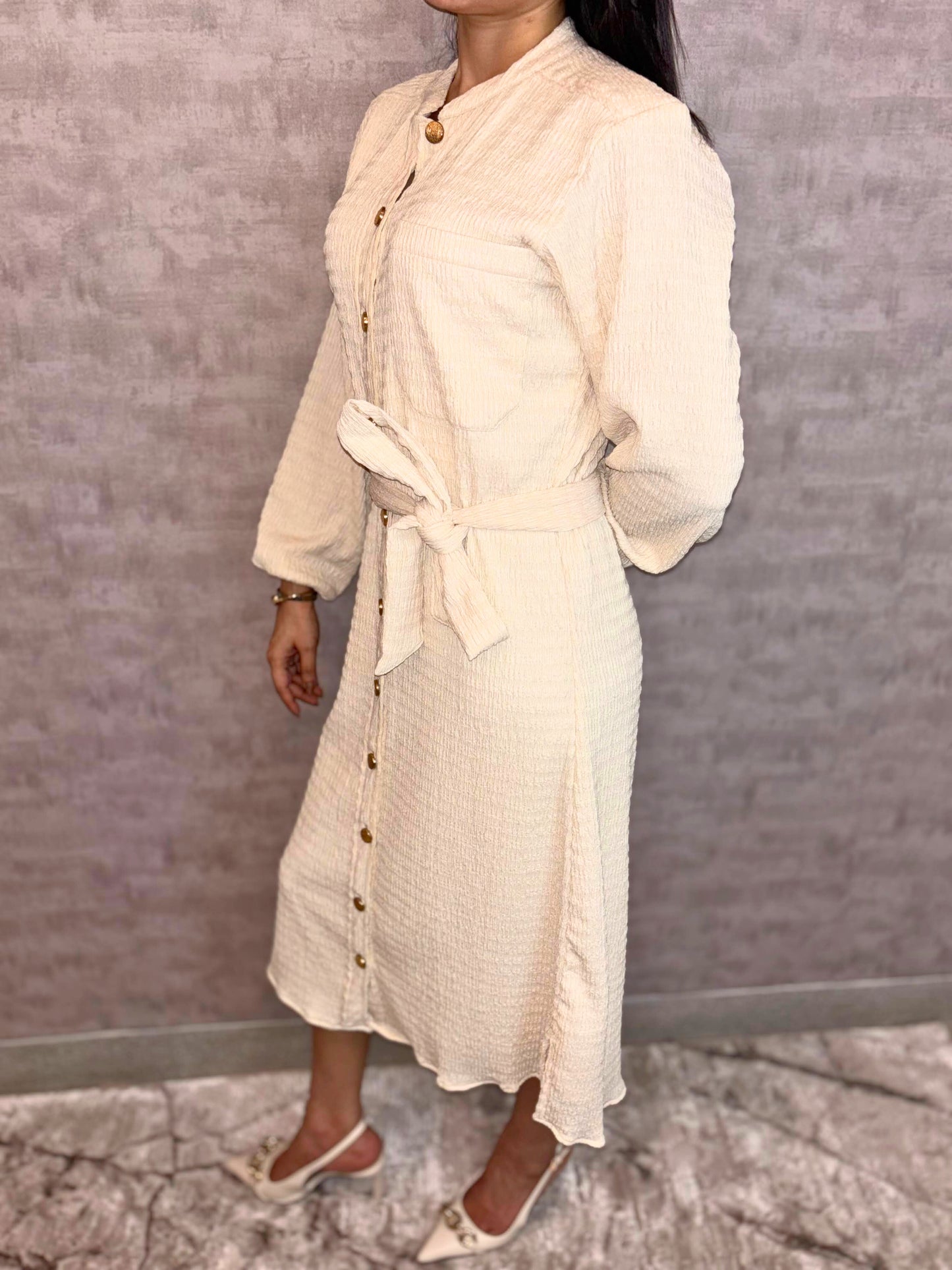 Experience versatile elegance with the Gilded Ivory Dress, a highlight of Taraashi's Platinum Collection. Crafted from uniquely textured fabric in ivory, this versatile piece blends everyday comfort with understated polish. Styled like a classic shirt dress with a defining tie belt, it's distinguished by a row of luminous gold buttons lending an elevated touch.