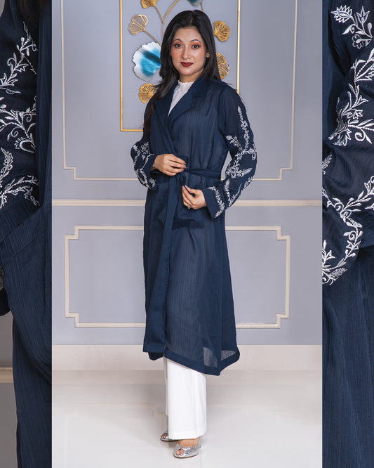 Woman wearing a navy blue embroidered abaya with white pants against a decorative wall.