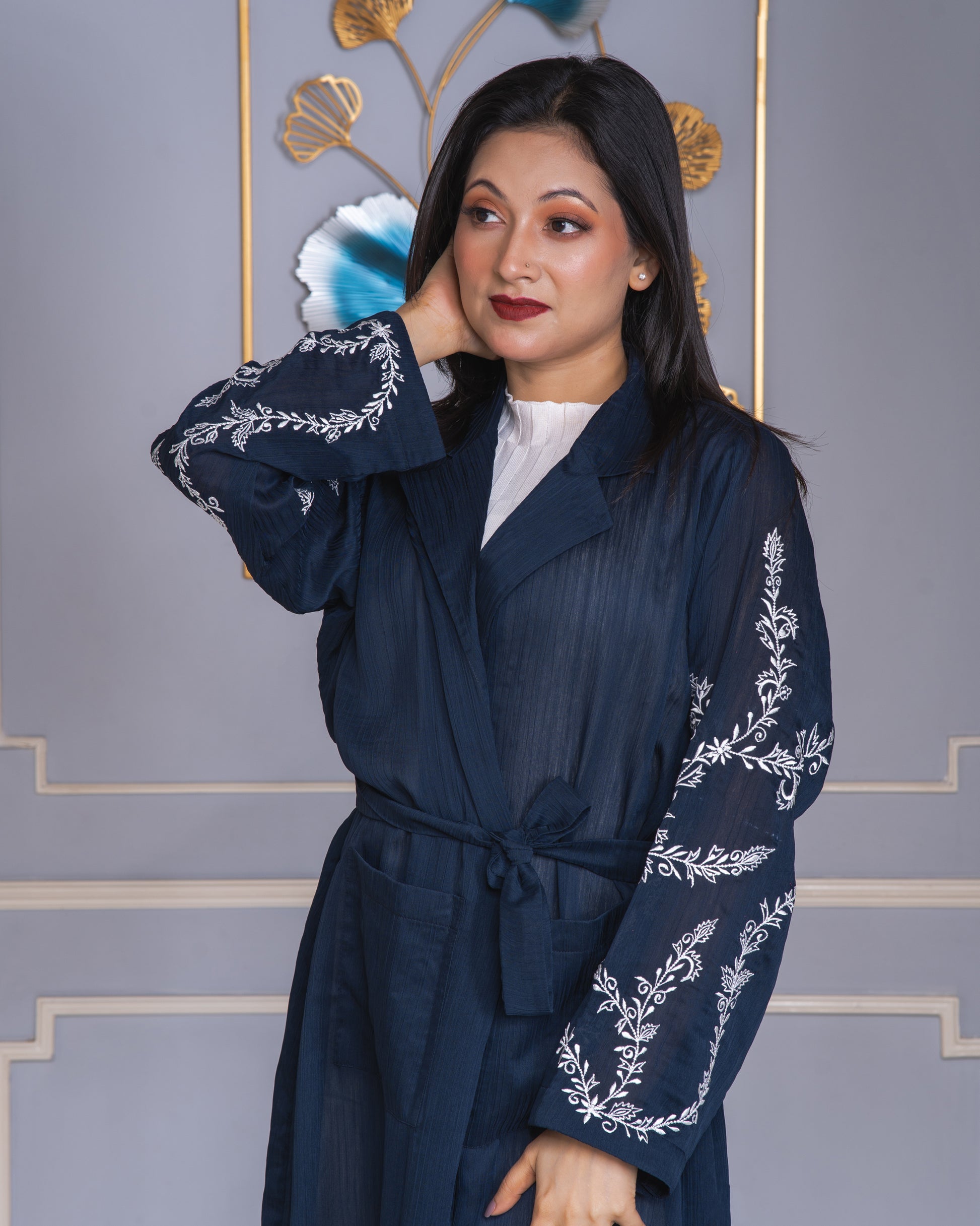 Woman wearing a dark blue embroidered coat against a decorative wall.